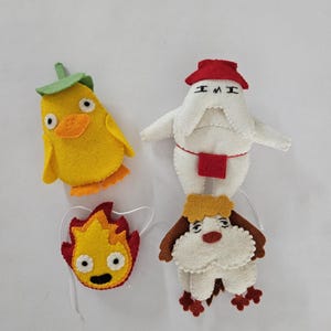 May include: Four handmade felt character ornaments. A yellow chick with a green leaf, a white figure with a red hat, a yellow and red flame, and a white dog with brown accents. Each has a string for hanging.