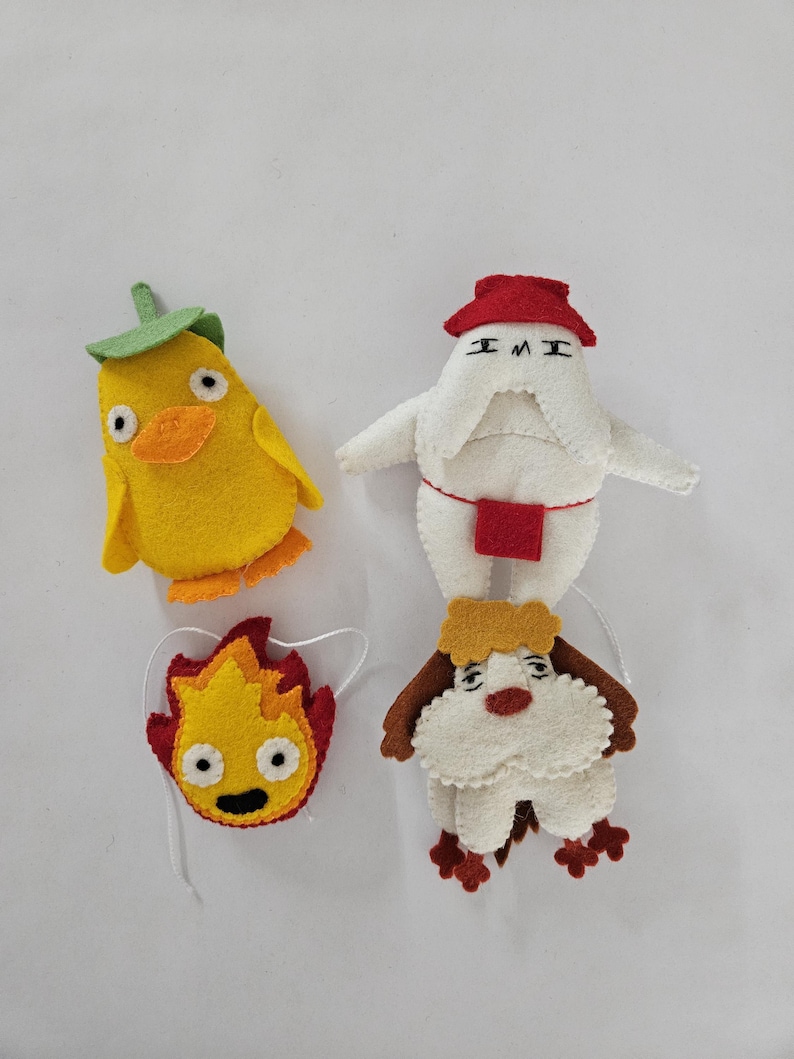 May include: Four handmade felt character ornaments. A yellow chick with a green leaf, a white figure with a red hat, a yellow and red flame, and a white dog with brown accents. Each has a string for hanging.