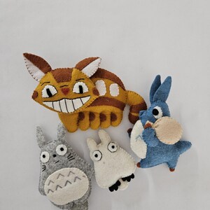 May include: A collection of handmade felt plush toys featuring characters from the animated film "My Neighbor Totoro." The toys include a large yellow cat-like creature, a gray Totoro, a white creature, and a blue creature.