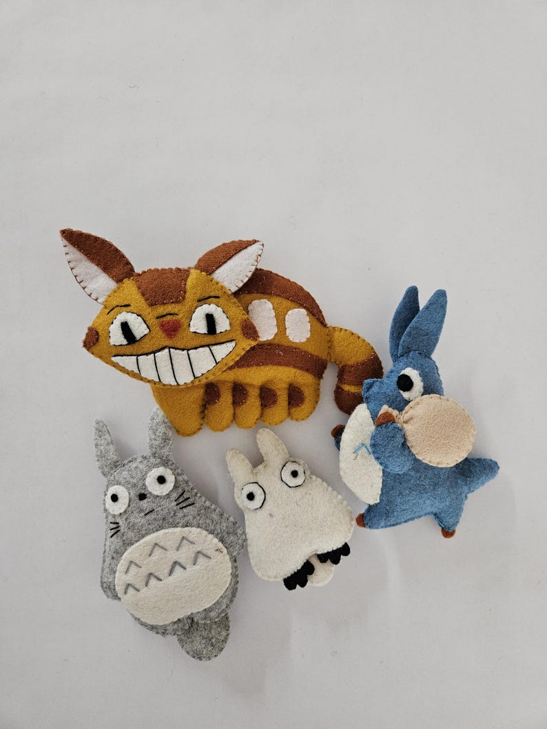 May include: A collection of handmade felt plush toys featuring characters from the animated film "My Neighbor Totoro." The toys include a large yellow cat-like creature, a gray Totoro, a white creature, and a blue creature.