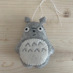 Op de afbeelding: Een grijs vilten Totoro-ornament met witte vilten accenten en een touwtje om op te hangen.