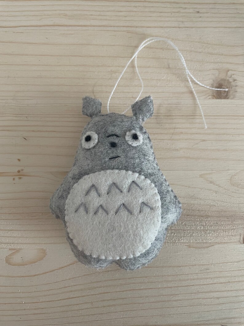 Op de afbeelding: Een grijs vilten Totoro-ornament met witte vilten accenten en een touwtje om op te hangen.