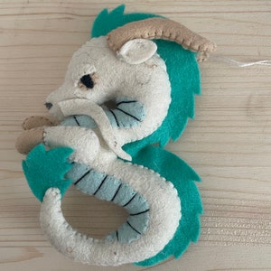 Op de afbeelding: Een witte vilt draak met turquoise groene accenten. De draak heeft een gekrulde staart en een klein hoorn op zijn kop.