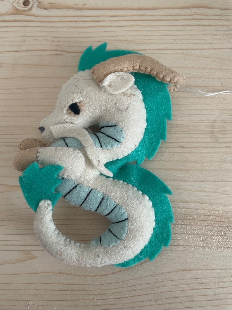 Op de afbeelding: Een witte vilt draak met turquoise groene accenten. De draak heeft een gekrulde staart en een klein hoorn op zijn kop.