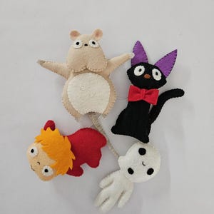 May include: A collection of handmade felt character figures. Includes a beige hippo, a black cat with a red bow tie and purple ears, a red and orange figure, and a white ghost-like figure. All figures have stitched details and are on a white background.