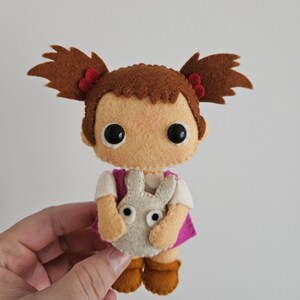 May include: A handmade felt doll depicting a girl with brown pigtails, each tied with a red bow. She is dressed in a white shirt, a pink vest, and holds a grey bunny. The doll has large black eyes and brown boots.