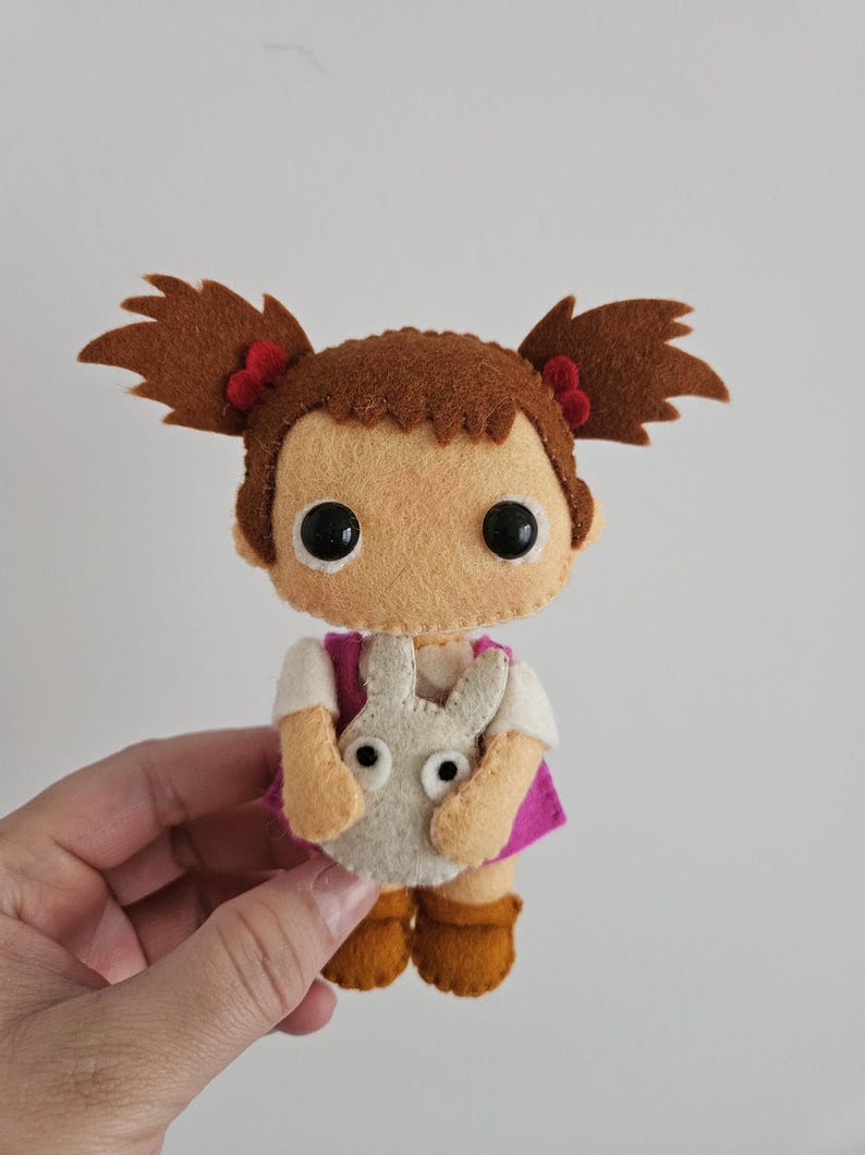 May include: A handmade felt doll depicting a girl with brown pigtails, each tied with a red bow. She is dressed in a white shirt, a pink vest, and holds a grey bunny. The doll has large black eyes and brown boots.