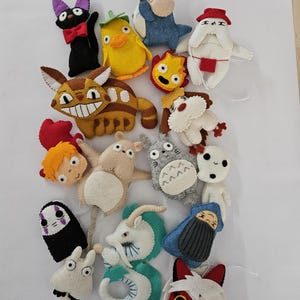 May include: A collection of colorful felt character plushies, including a black cat with a red bow tie, a yellow duck, and a blue rabbit. Other characters include a brown and tan creature with a large grin, and a gray creature with a white belly.