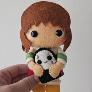 May include: A handmade felt doll of a girl with brown hair, wearing a white shirt with green stripes, a red skirt, and yellow shoes. The doll is holding a small black and white creature with purple accents.