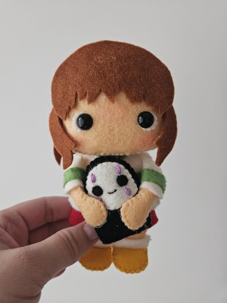 May include: A handmade felt doll of a girl with brown hair, wearing a white shirt with green stripes, a red skirt, and yellow shoes. The doll is holding a small black and white creature with purple accents.