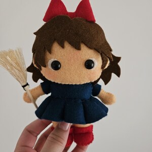May include: A handmade felt doll of a young girl with brown hair, a red bow, and a navy blue dress. The doll is holding a small broom and wearing red boots. The doll's face is light tan with black eyes.