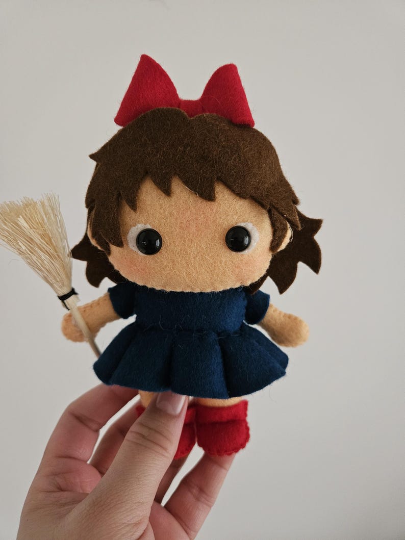 May include: A handmade felt doll of a young girl with brown hair, a red bow, and a navy blue dress. The doll is holding a small broom and wearing red boots. The doll's face is light tan with black eyes.