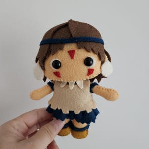 May include: A handmade felt doll with brown hair, a blue headband, and a tan face. The doll wears a tan top with a blue skirt and yellow shoes. Red markings are on the doll's face. The doll is held by a hand.