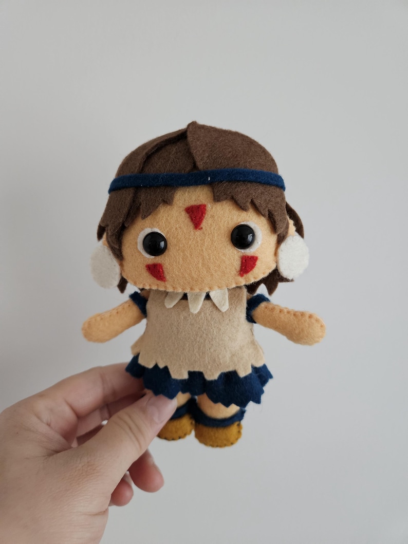 May include: A handmade felt doll with brown hair, a blue headband, and a tan face. The doll wears a tan top with a blue skirt and yellow shoes. Red markings are on the doll's face. The doll is held by a hand.