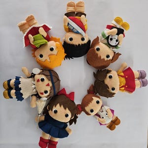 May include: A circular arrangement of handmade felt dolls. Each doll features a distinct outfit and hairstyle, with details such as bows, hats, and accessories. The dolls are in various colours, including red, blue, yellow, and brown.
