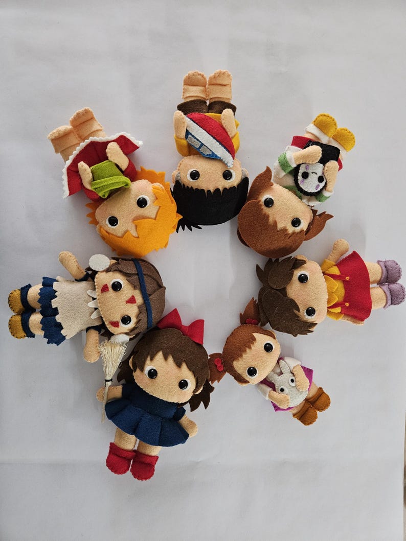 May include: A circular arrangement of handmade felt dolls. Each doll features a distinct outfit and hairstyle, with details such as bows, hats, and accessories. The dolls are in various colours, including red, blue, yellow, and brown.