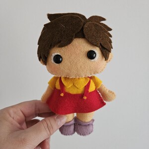 May include: A handmade felt doll with brown hair, large black eyes, and a tan face. The doll wears a yellow top with a red skirt and purple shoes. The doll is held by a hand against a white background.