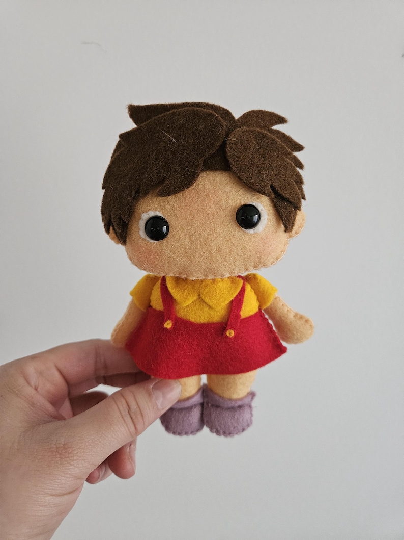 May include: A handmade felt doll with brown hair, large black eyes, and a tan face. The doll wears a yellow top with a red skirt and purple shoes. The doll is held by a hand against a white background.
