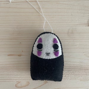 Op de afbeelding: Een wit vilten ornament met een zwart gezicht en paarse ogen. Het ornament is gevormd als een persoon en heeft een touwtje om op te hangen.