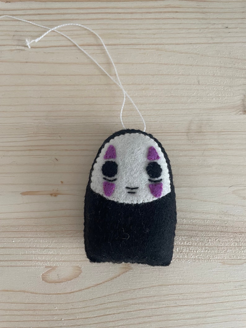 Op de afbeelding: Een wit vilten ornament met een zwart gezicht en paarse ogen. Het ornament is gevormd als een persoon en heeft een touwtje om op te hangen.