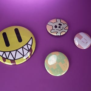 May include: Four round pinback buttons with various designs on a purple background. One button features a yellow smiley face with a menacing grin and blood-like drips. Other buttons have a skull bunny, a geometric pattern, and a green and yellow design.