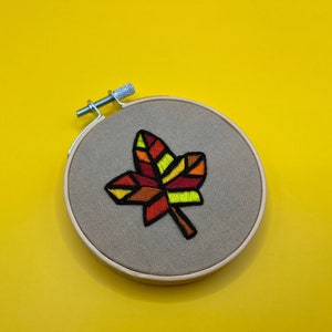 Pode incluir: Um aro de bordado marrom com um design de folha geométrico multicolorido. A folha é feita com fio vermelho, laranja, amarelo e verde.