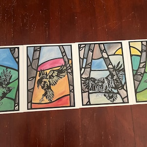 May include: Four framed art prints featuring stylized ravens in flight, set against colorful stained-glass window backgrounds. Each print showcases a different pose of the raven, with vibrant hues of blue, green, orange, and pink. The artwork is set on a dark wood surface.