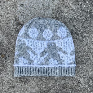 May include: A gray and white knit beanie hat. The hat features a repeating pattern of white figures on a gray background. The brim is a solid gray color. The hat is made of a soft, textured material.