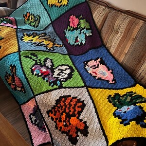 Puede incluir: Una manta de ganchillo hecha a mano con varios dise&ntilde;os de personajes pixelados de colores. La manta est&aacute; hecha de cuadrados en tonos de azul, verde, amarillo, morado y rosa. La manta est&aacute; sobre un sof&aacute; marr&oacute;n.