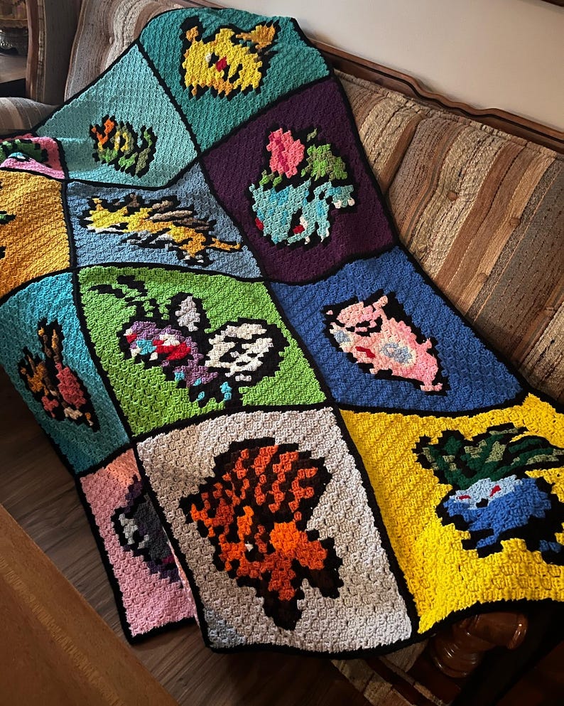 Puede incluir: Una manta de ganchillo hecha a mano con varios dise&ntilde;os de personajes pixelados de colores. La manta est&aacute; hecha de cuadrados en tonos de azul, verde, amarillo, morado y rosa. La manta est&aacute; sobre un sof&aacute; marr&oacute;n.