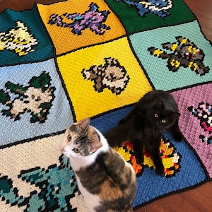 Puede incluir: Una manta de ganchillo pixelada y hecha a mano con varios dise&ntilde;os de personajes coloridos. La manta est&aacute; dividida en cuadrados de diferentes colores, cada uno mostrando un personaje &uacute;nico. Dos gatos descansan sobre la manta.