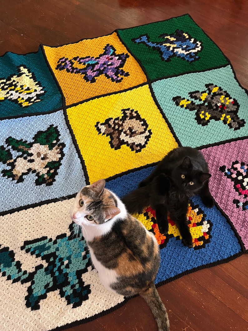 Puede incluir: Una manta de ganchillo pixelada y hecha a mano con varios dise&ntilde;os de personajes coloridos. La manta est&aacute; dividida en cuadrados de diferentes colores, cada uno mostrando un personaje &uacute;nico. Dos gatos descansan sobre la manta.