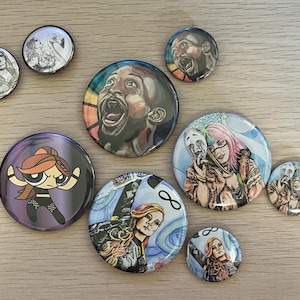 May include: A collection of round pinback buttons with various designs. Some buttons feature illustrations of people, including a basketball player and a cartoon character. Other buttons have drawings of women.