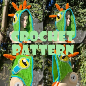 May include: Green crochet hooded hat with orange and blue accents. The hat features a large eye with a yellow iris and blue pupil. The hat is crocheted in a textured stitch and has a long, flowing hood.