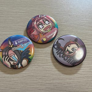 May include: Three colorful anime-style button pins with different character designs and text. One pin says "Awful Mood Down", another says "Umazed!", and the third says "Bakushin". The pins are round and have a glossy finish.