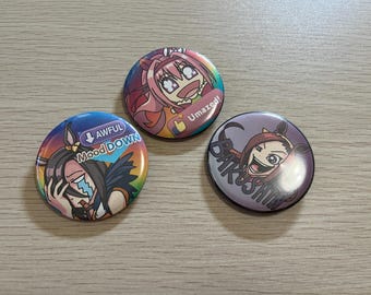 Könnte beinhalten: Drei bunte Ansteckbuttons im Anime-Stil mit verschiedenen Charakterdesigns und Texten. Einer sagt "Awful Mood Down", ein anderer "Umazed!" und der dritte "Bakushin". Die Buttons sind rund und haben ein glänzendes Finish.