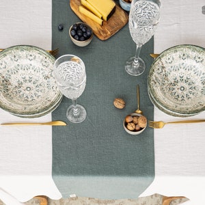 Könnte beinhalten: Ein grüner Tischläufer aus Leinen auf weißem Hintergrund. Zwei Gedecke sind mit grün-weißen gemusterten Schalen, goldenem Besteck und klaren Gläsern gedeckt. Eine kleine Schale mit Nüssen befindet sich in der Mitte des Läufers.