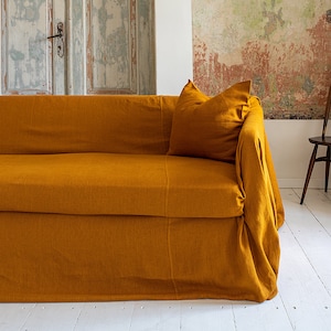 May include: A mustard yellow linen sofa cover with a matching pillow. The sofa cover has a gathered skirt and is draped over a sofa.