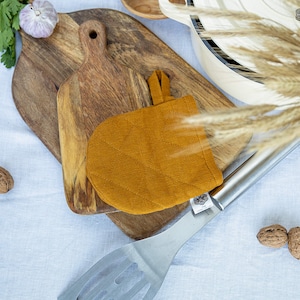 Può includere: Un guanto da forno in lino giallo senape con un'ansa a forma di anello. Il guanto da forno è sopra un tagliere di legno. Il tagliere è sopra una tovaglia di lino bianca. Il testo "THE LINEN VALLEY" è visibile nell'angolo in alto a destra dell'immagine.
