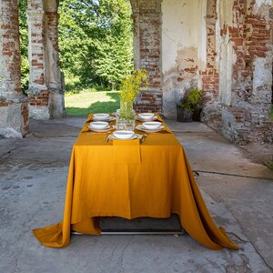 May include: A mustard yellow linen tablecloth set for a dinner party with white plates and silverware. The table is set in a rustic outdoor setting with brick columns and greenery in the background.