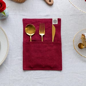 May include: A burgundy linen cutlery holder with gold-colored spoon, fork, and knife. The utensils are neatly arranged in the holder, which has a small tag with text. The holder is on a white tablecloth, with a plate and a flower in the background.