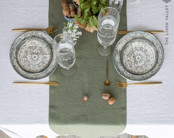 May include: A green linen table runner with two place settings. Each place setting includes a white and blue patterned dinner plate, a gold fork and knife, and a glass of water. There are also nuts and a small plant on the table runner.