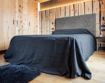 May include: A bedroom with a wooden wall and a large bed with a black linen duvet. The bed has a grey headboard and a wooden bedside table with a cup of coffee on it.
