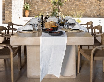 May include: A concrete table with a white linen tablecloth set for a dinner party. The table is surrounded by wooden chairs and there are plates, glasses, and cutlery set on the table. There is a bowl of fruit in the centre of the table.