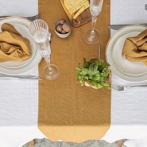 May include: A white tablecloth with a mustard yellow linen runner down the center. Two place settings are visible, each with a white plate, a gold napkin, and a clear glass. A small plant in a brown paper bag is in the center of the runner.
