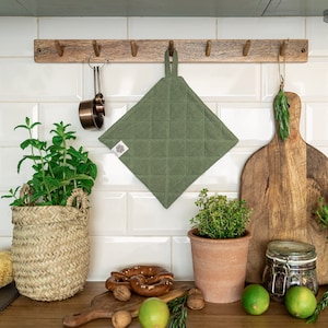 Puede incluir: Un agarrador acolchado verde oliva colgado de un estante de madera en una cocina. El agarrador tiene forma cuadrada con un lazo para colgar. Otros elementos de cocina incluyen una tabla de cortar, tazas medidoras y hierbas en macetas.