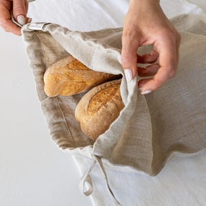 Könnte beinhalten: Ein beiger Brotbeutel aus Leinen enthält zwei knusprige Brote. Der Beutel ist offen und zeigt die goldbraunen Laibe. Der Beutel hat einen Kordelzugverschluss und ein kleines Etikett. Der Hintergrund ist eine weiße Oberfläche.