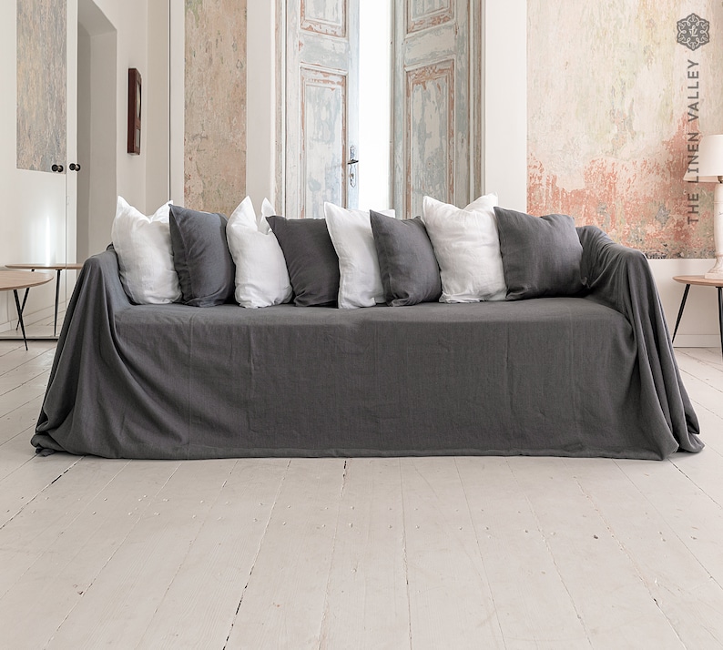 May include: A dark gray sofa cover draped over a couch, featuring a series of alternating white and dark gray square throw pillows arranged along the backrest. The fabric has a visible texture and hangs down to the floor on both sides of the sofa.