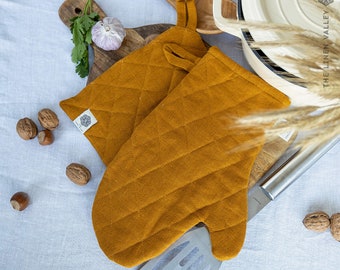 May include: A pair of mustard yellow oven mitts featuring a quilted diamond pattern and a small fabric tag with the text THE LINEN VALLEY. Each mitt includes a fabric loop for hanging, constructed from a textured fabric with visible stitching.