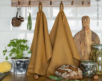 May include: Two mustard yellow kitchen towels with hanging loops, suspended from a wooden wall rack. The towels feature a visible woven texture and are displayed in a kitchen setting alongside a loaf of bread, herbs, and wooden cutting board.